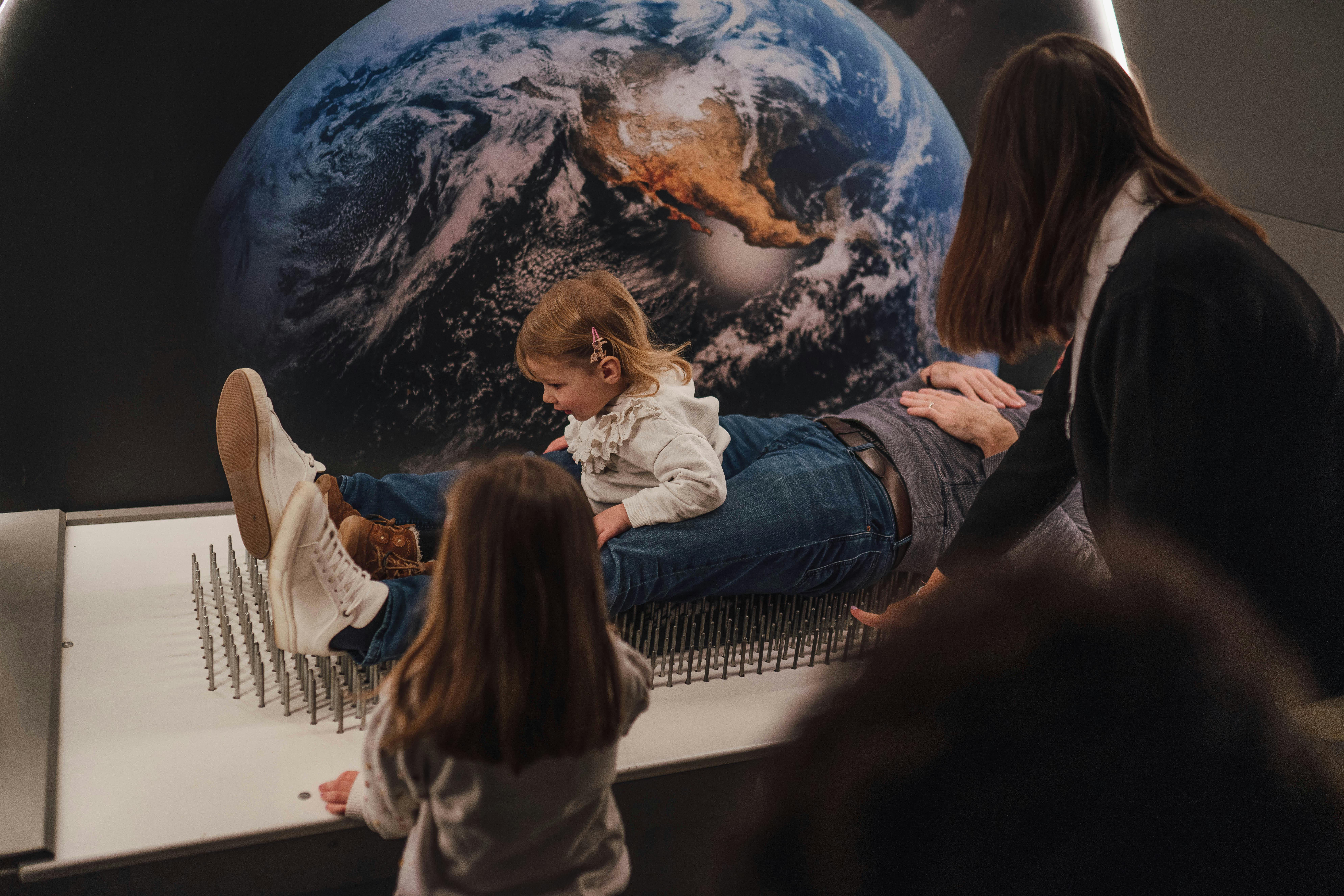 Un enfant est assis sur un adulte allongé sur un lit de clous, avec la Terre en arrière-plan. Une fillette observe à proximité.