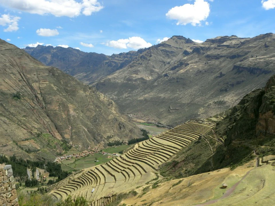 Terraced farmland on a mountain slope with a village in a valley, surrounded by high mountains and a partly cloudy sky.