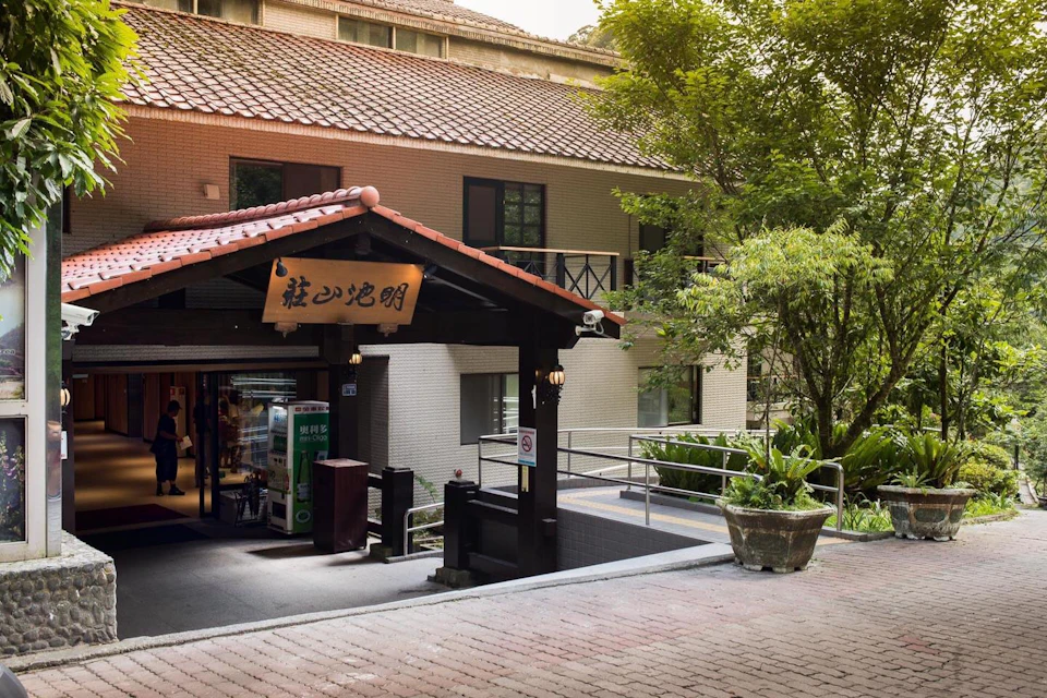 A building entrance with a tiled roof and wooden frame, surrounded by greenery, with a sign featuring Chinese characters.
