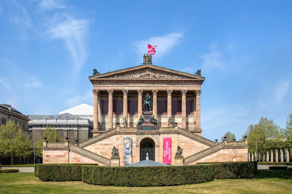 A neoclassical building with columns, a statue in front, and a red banner displayed, under a clear blue sky.
