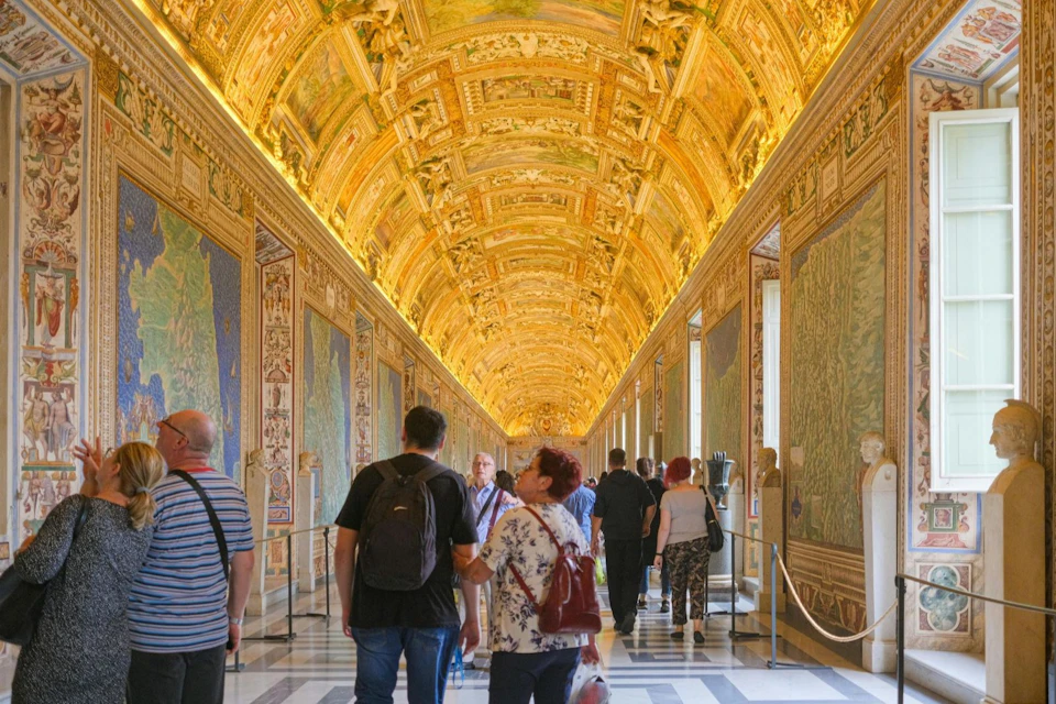 People walking through an ornate hallway with a decorated golden ceiling and detailed wall art.