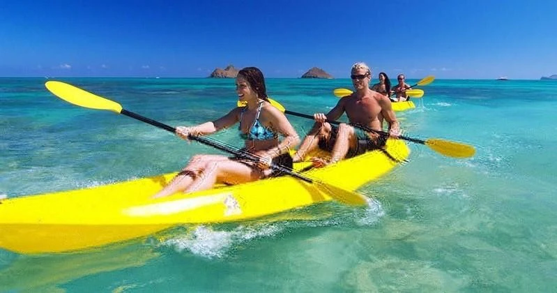 Two people kayaking on a yellow kayak in clear turquoise water with small islands visible in the background under a blue sky.