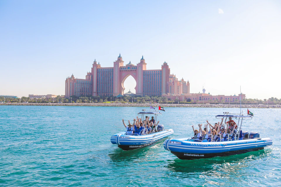 Two tour boats with people waving on a turquoise sea, with a large pink hotel with a central archway in the background.