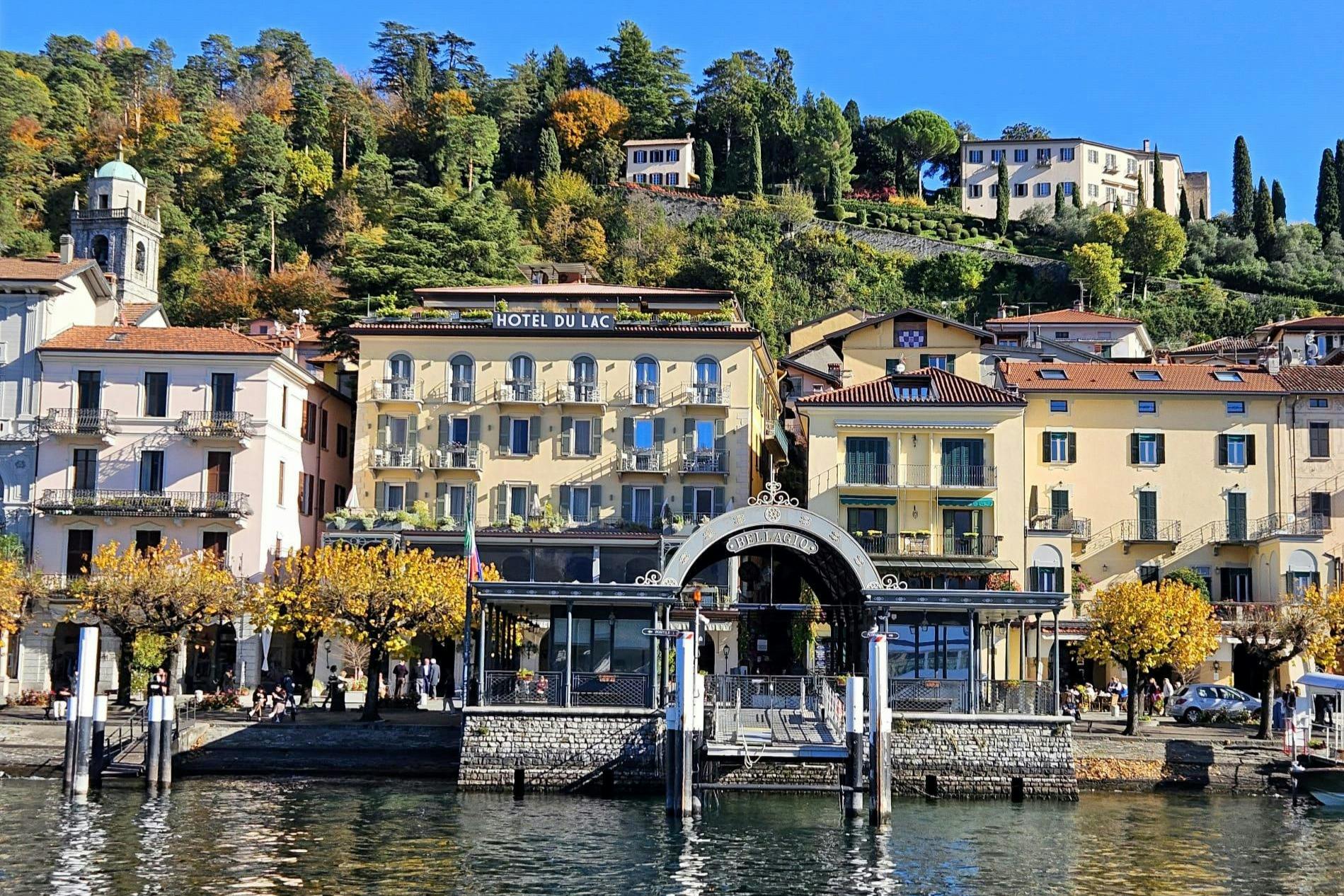 A lakeside hotel with yellow buildings and a small dock, set against a hillside with green trees and several houses.