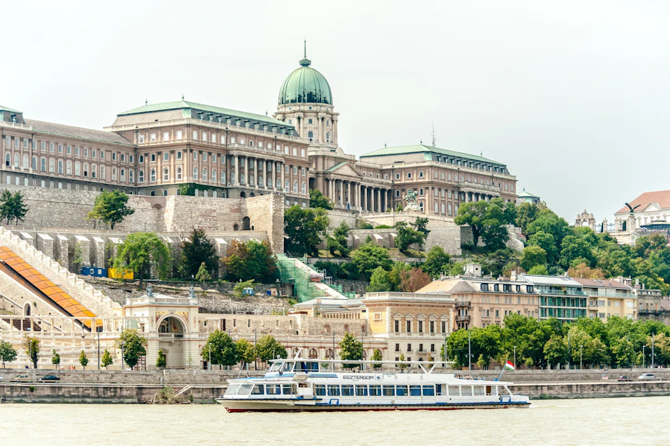 A large historic building with a dome on a hill, with a boat on a river in the foreground and trees around.