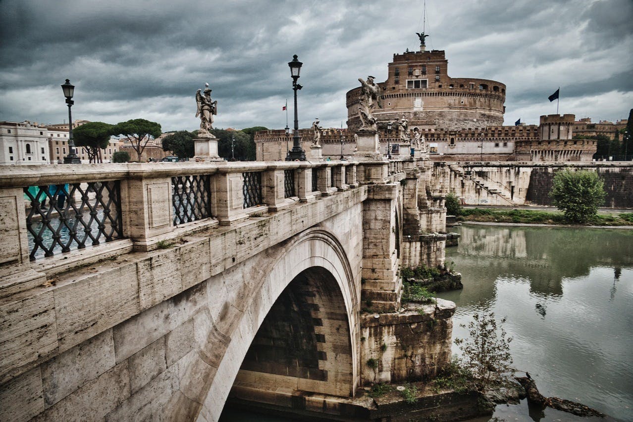 Stone bridge with statues and lamp posts crossing a river, leading to a large, cylindrical fortress under a cloudy sky.