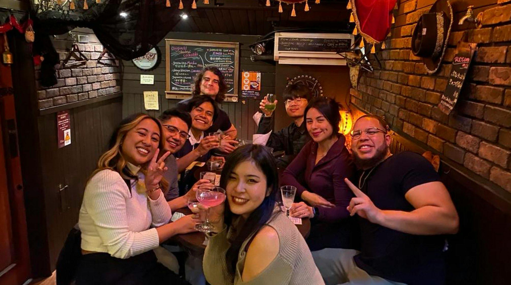 A group of eight people sitting together at a table in a cozy indoor setting, smiling and holding drinks.