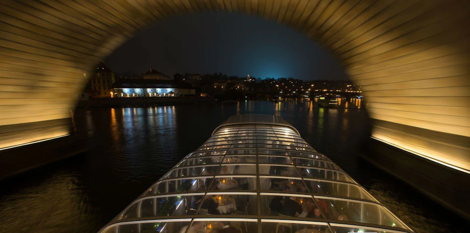 A glass-roofed boat with dining tables inside on a river at night, with lit buildings and reflections visible in the background.