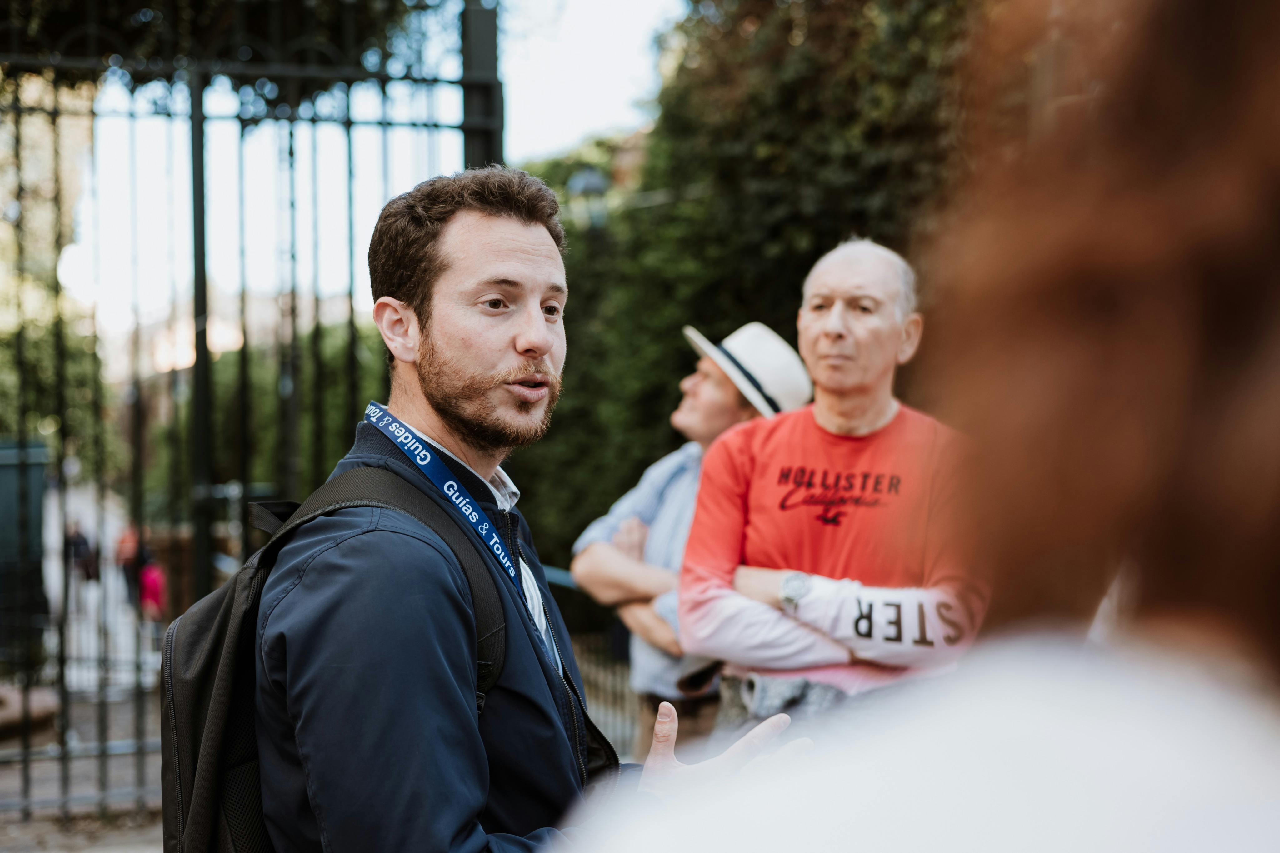 Prohlídka s průvodcem promlouvá ke skupině lidí venku. Jeden muž v červené košili a druhý v klobouku pozorně naslouchá v pozadí.