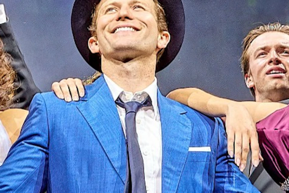 A group of performers in formal attire stand on a lit stage holding hats, with theatrical lighting and set design in the background.