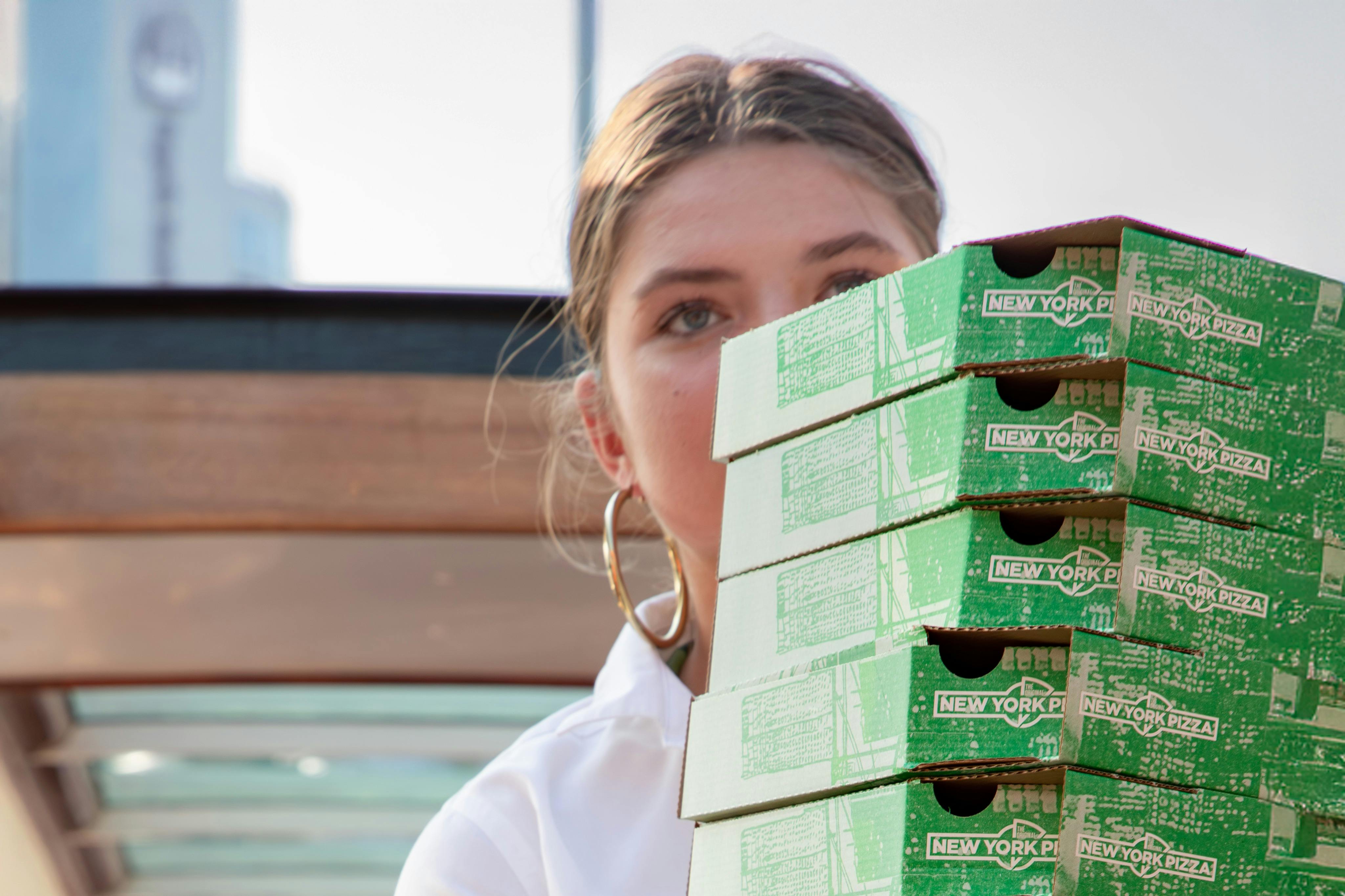 초록색 "뉴욕 피자" 상자를 들고 있는 사람으로, 코에서 위로 보이며, 실외 배경이 흐릿합니다.