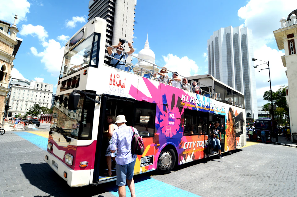 Tourists boarding and seated on a colorful, double-decker city tour bus on a sunny day with modern buildings in the background.