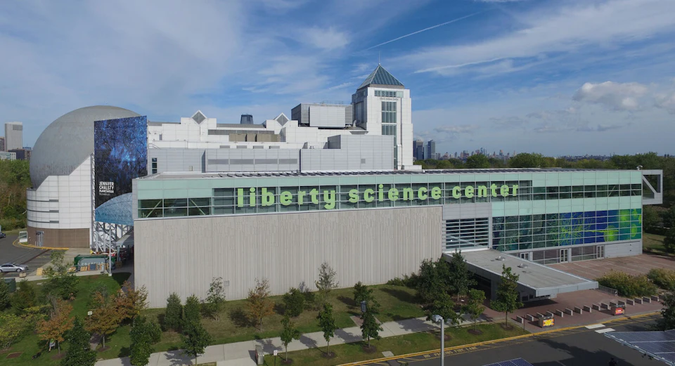 Modern building with "Liberty Science Center" written on the facade, surrounded by greenery, under a blue sky.