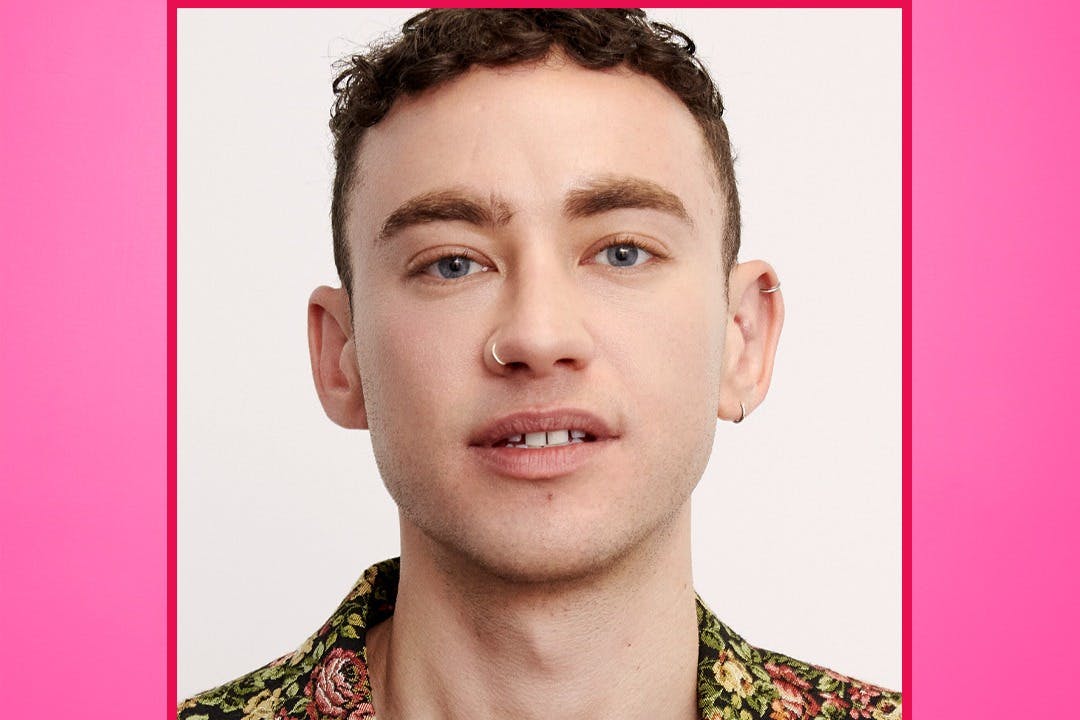 Ένα άτομο με κοντά σγουρά μαλλιά, που φοράει ένα φλοράλ πουκάμισο, έχει κρίκο στη μύτη και σκουλαρίκι στο αυτί, σε απλό φόντο.