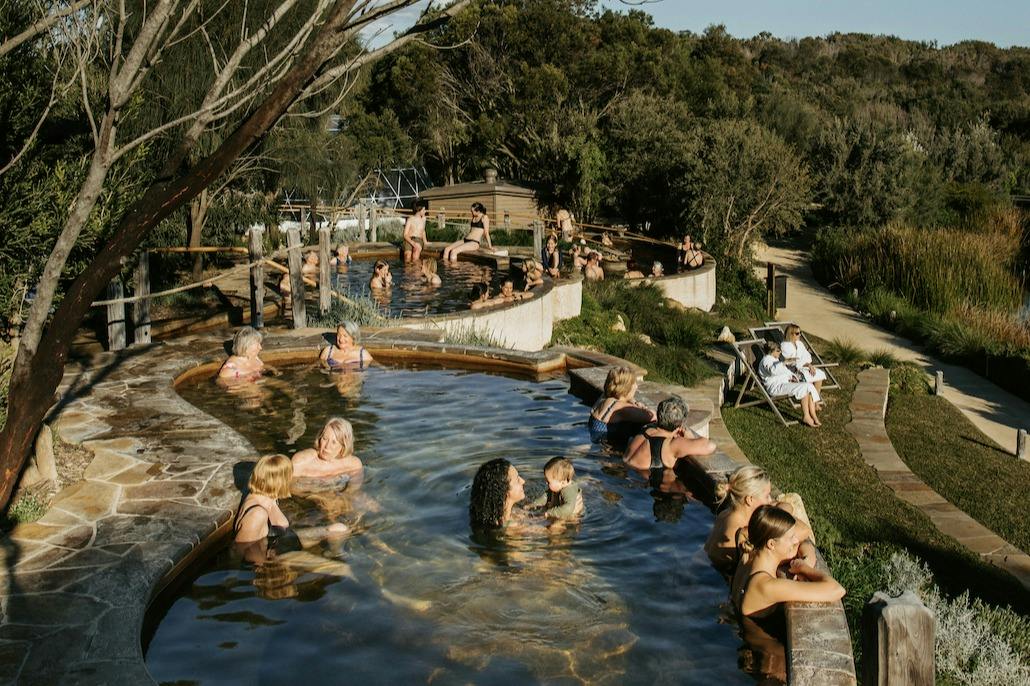People enjoying outdoor hot spring pools surrounded by trees and a natural setting on a sunny day.