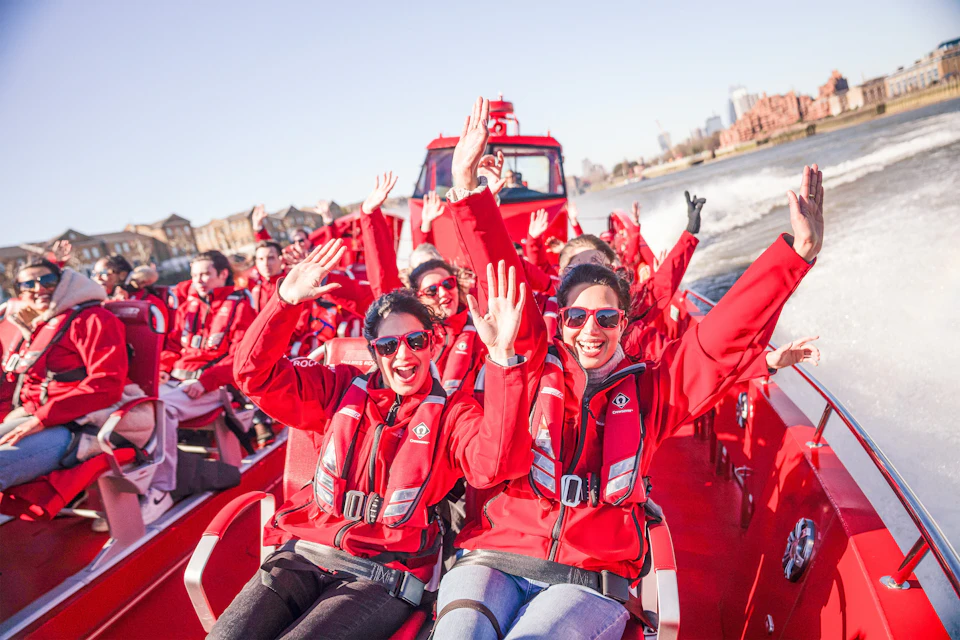 A group of people wearing red jackets and life vests enjoy a boat ride, smiling and waving, with a cityscape in the background.