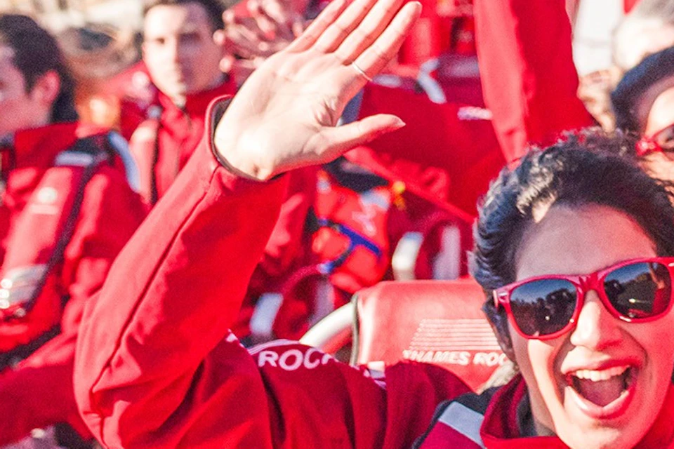 A group of people wearing red jackets and life vests enjoy a boat ride, smiling and waving, with a cityscape in the background.