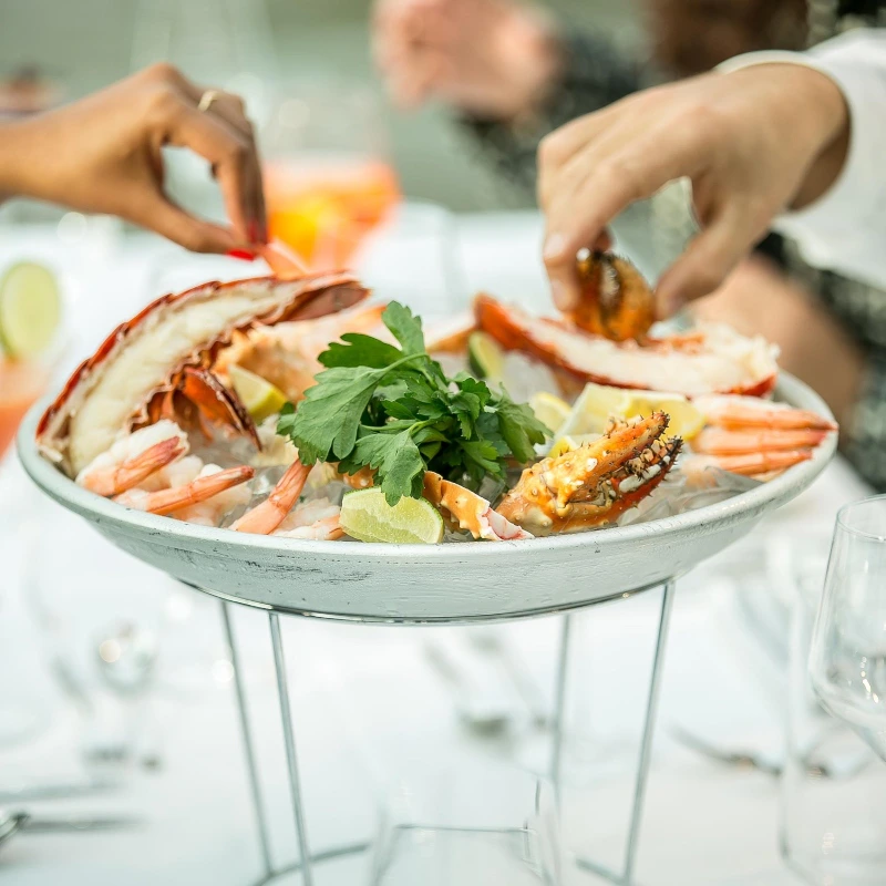 A seafood platter with lobster, shrimp, and garnishes, as two people reach to serve themselves at a dining table.