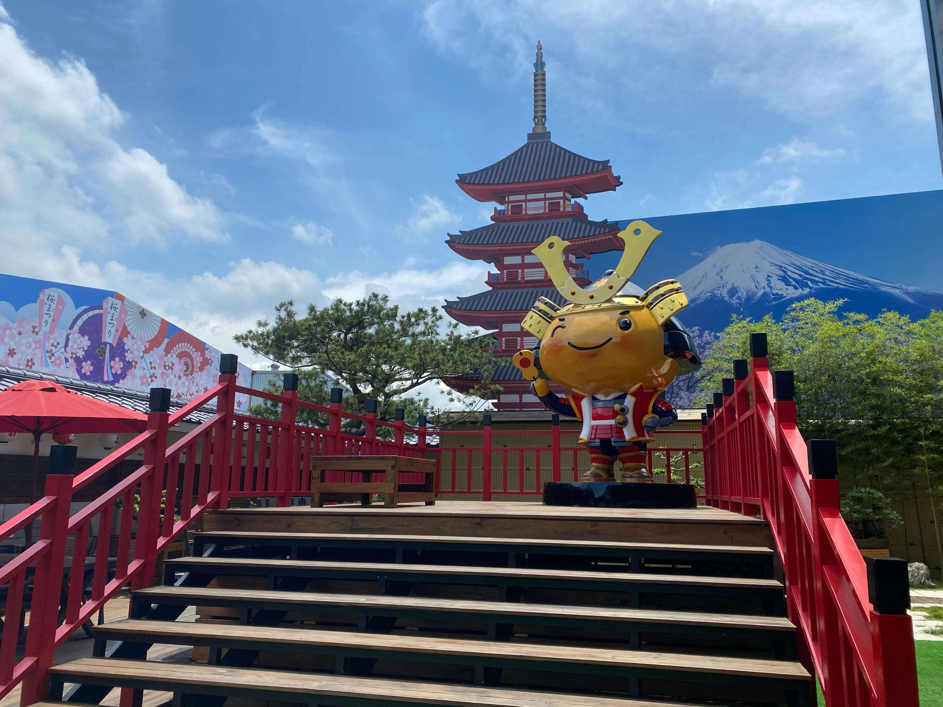 A colorful samurai-themed mascot stands on wooden steps near a red pagoda and a scenic background with Mount Fuji.