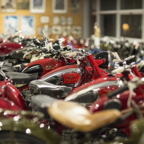 File di classiche Moto Guzzi rosse parcheggiate all'interno, con foto incorniciate sul muro sullo sfondo.