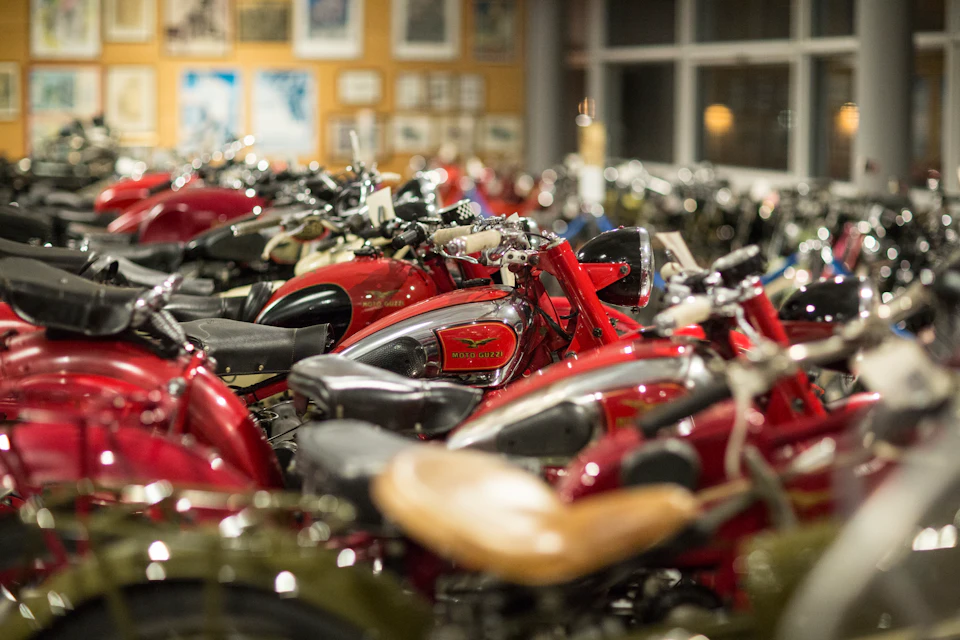Rows of classic red Moto Guzzi motorcycles tightly parked indoors, with framed pictures on the wall in the background.