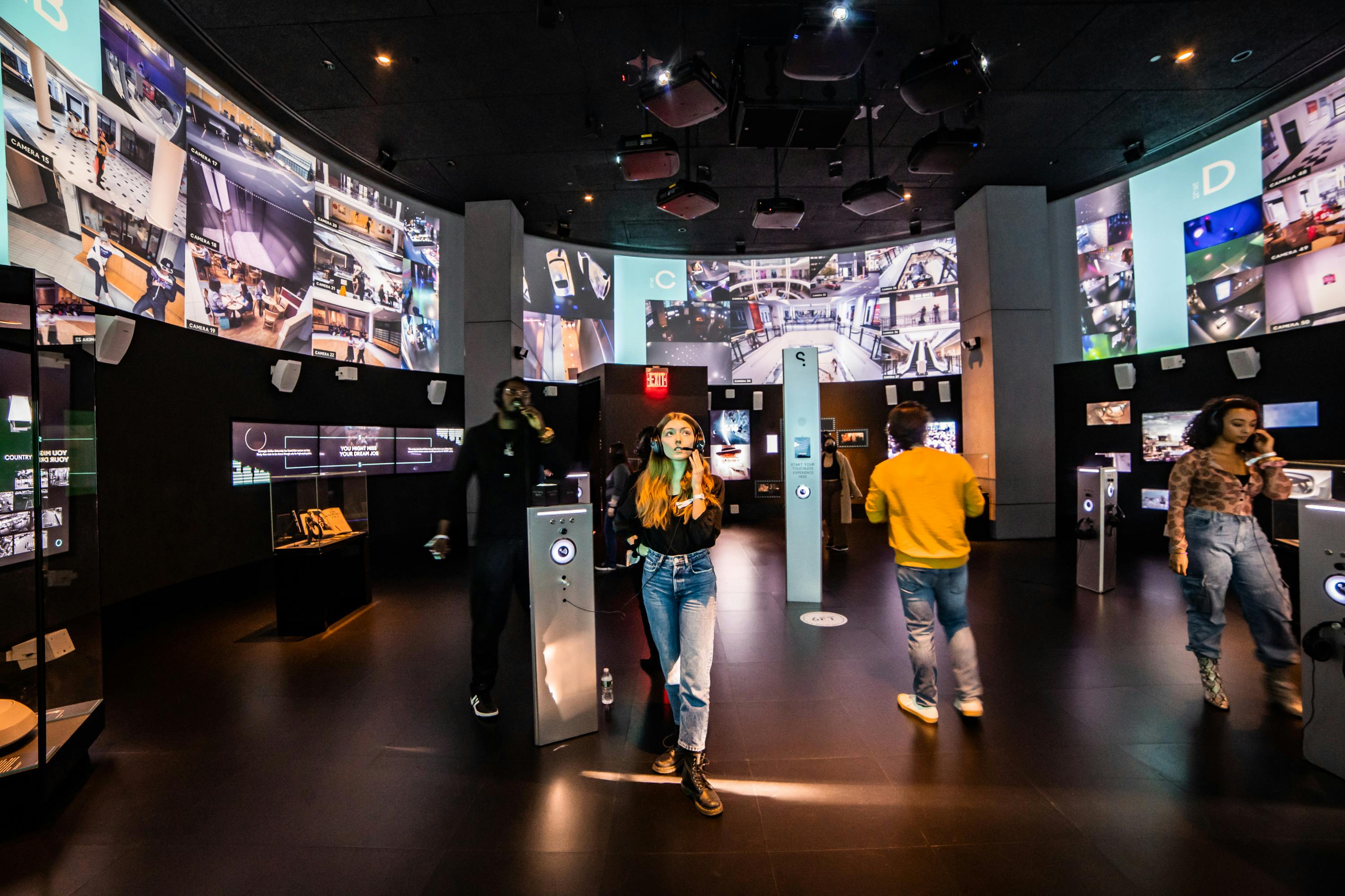Οι άνθρωποι αλληλεπιδρούν με σταθμούς ήχου σε ένα σκοτεινό δωμάτιο με πολλές μεγάλες οθόνες που εμφανίζουν διάφορες εικόνες.