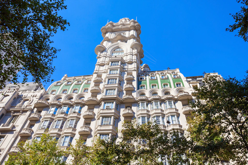 Tall ornate building with rounded balconies, green architectural accents, set against a clear blue sky, partially framed by trees.