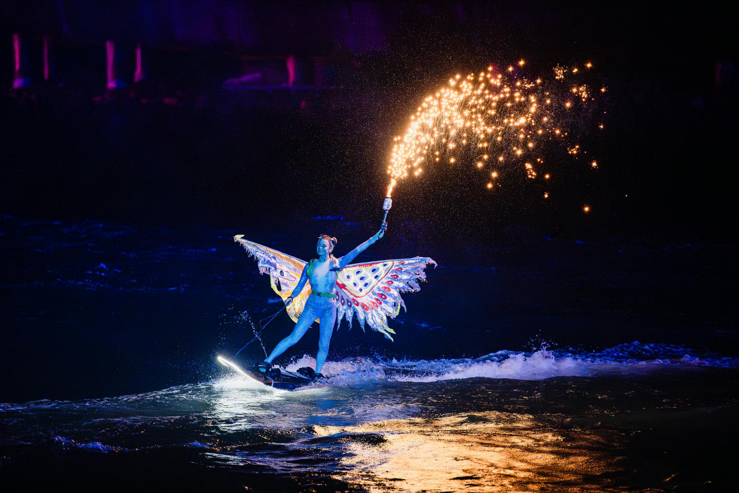 A performer in colorful butterfly wings water skiing at night, holding a sparkler emitting bright sparks.