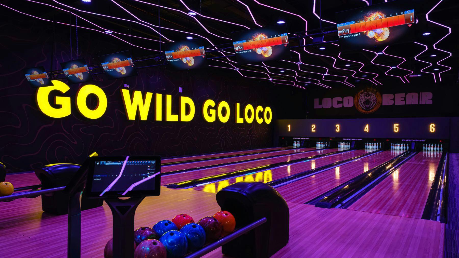 Μια αίθουσα μπόουλινγκ με φώτα νέον, πολύχρωμες μπάλες μπόουλινγκ, πολλαπλές λωρίδες και οθόνες που εμφανίζουν βαθμολογίες παραπάνω.