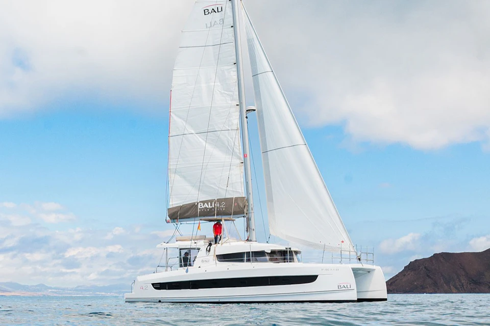 A white catamaran with large sails and a person in a red jacket on the deck, sailing on calm water under a cloudy sky.