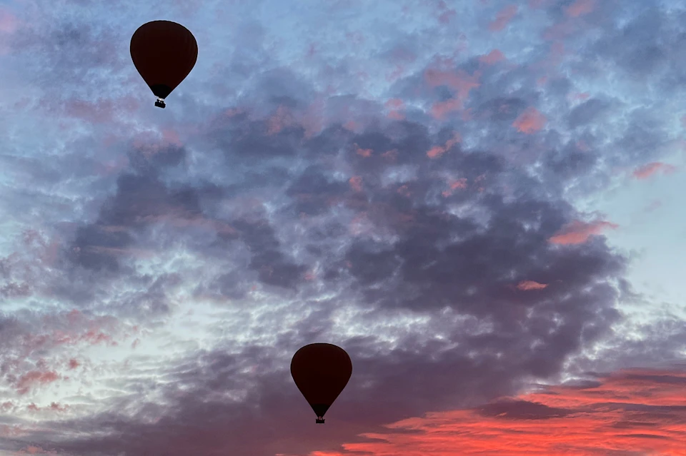 Two hot air balloons silhouetted against a colorful sky during sunrise or sunset, with clouds tinged in pink and purple hues.