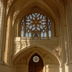 Interiör av en katedral med utsmyckade glasmålningar, välvda tak och stora trädörrar. Solljus strömmar genom fönster.