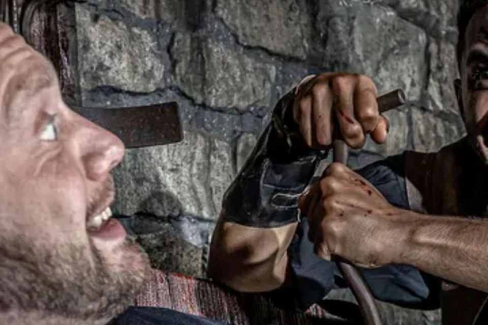 A man with dark makeup menacingly approaches another man sitting against a stone wall, holding a piece of curved metal.