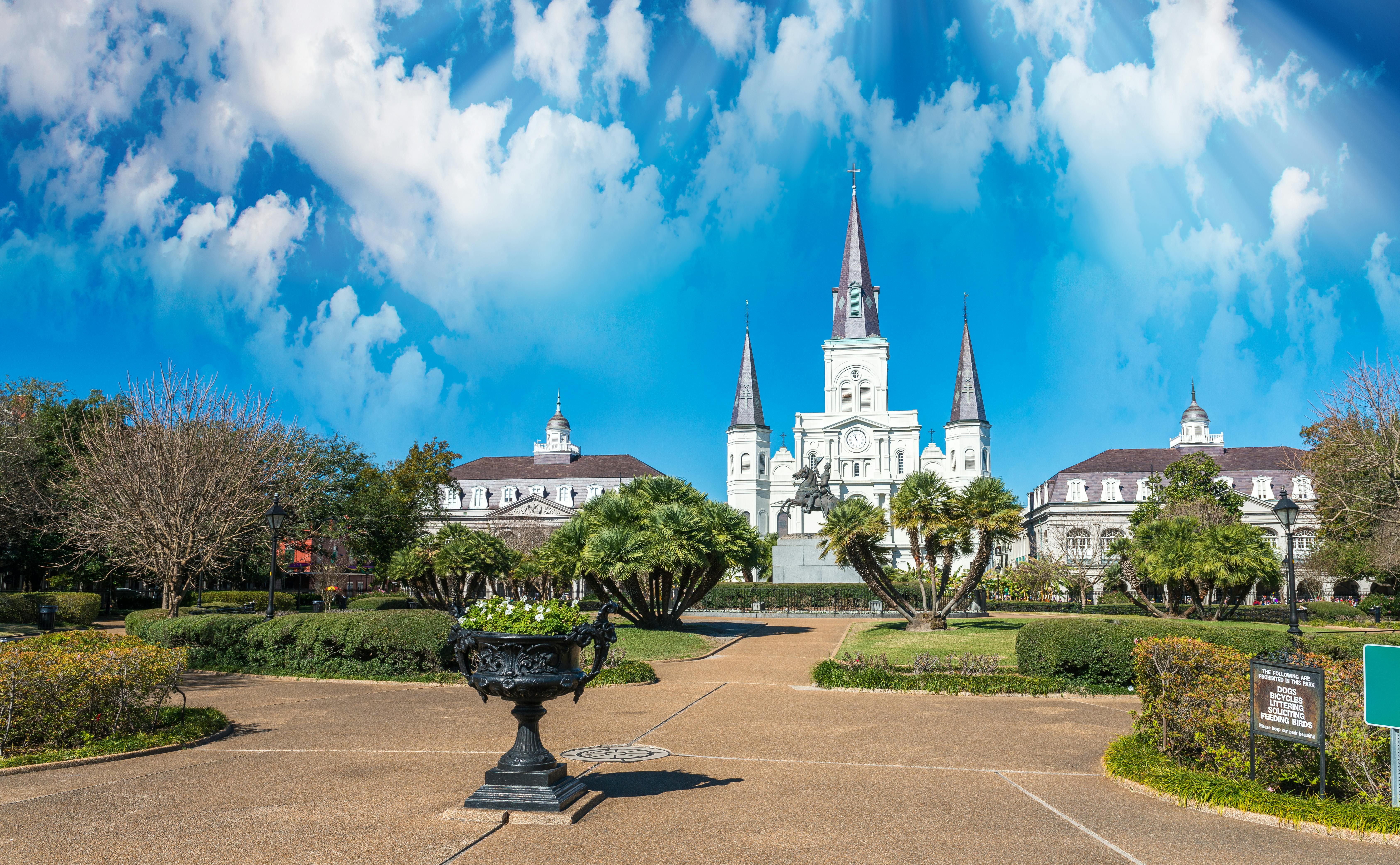 Park with neatly trimmed bushes and a statue in the center, flanked by historic buildings and a church with tall spires under a blue sky.