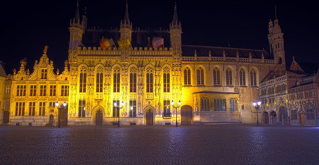 Illuminated Gothic-style buildings on a cobblestone square at night, with street lamps casting a warm glow.