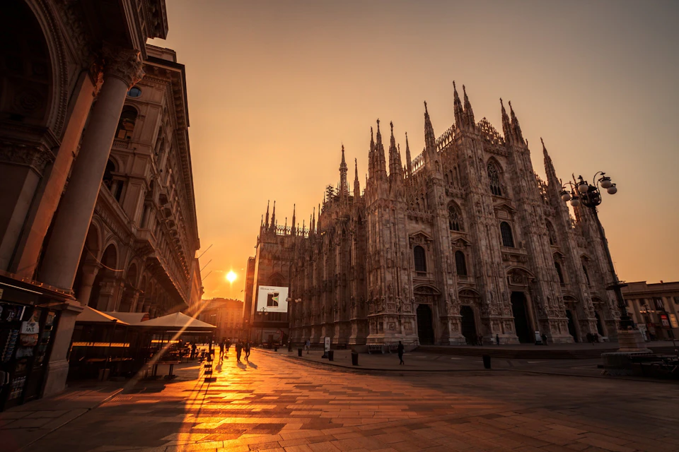 A historic cathedral and surrounding buildings illuminated by a warm sunset, with people walking in the square.