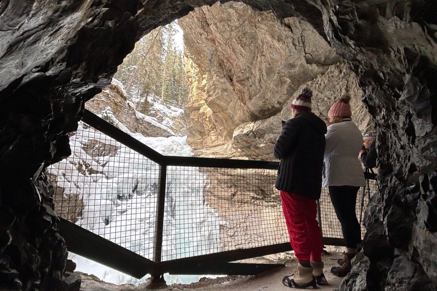 Two people in winter clothing stand in a cave, looking out at a snowy landscape with a frozen waterfall.