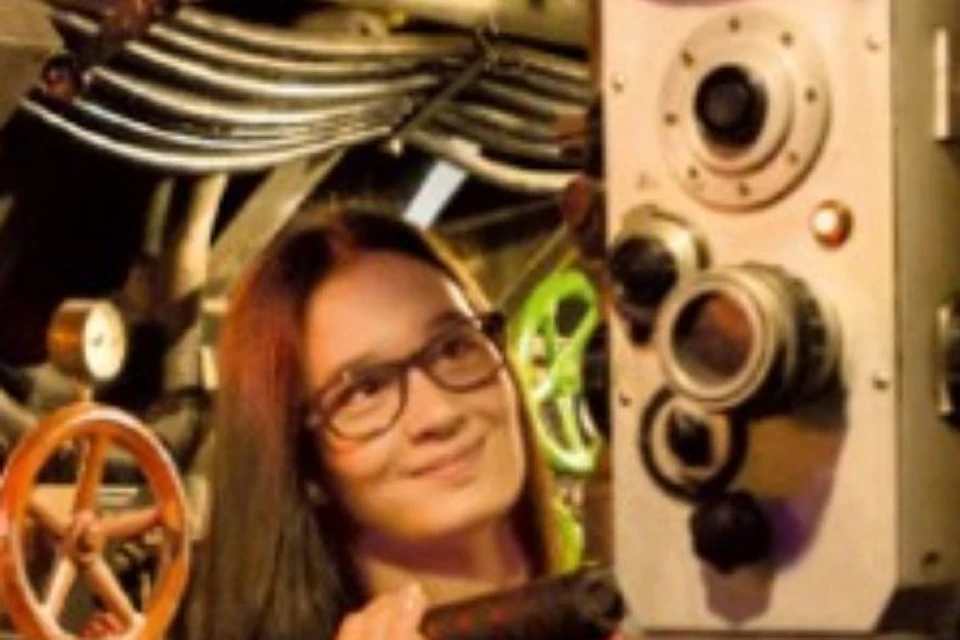 Three people standing inside a submarine control room, surrounded by dials and machinery, smiling and engaged with the equipment.