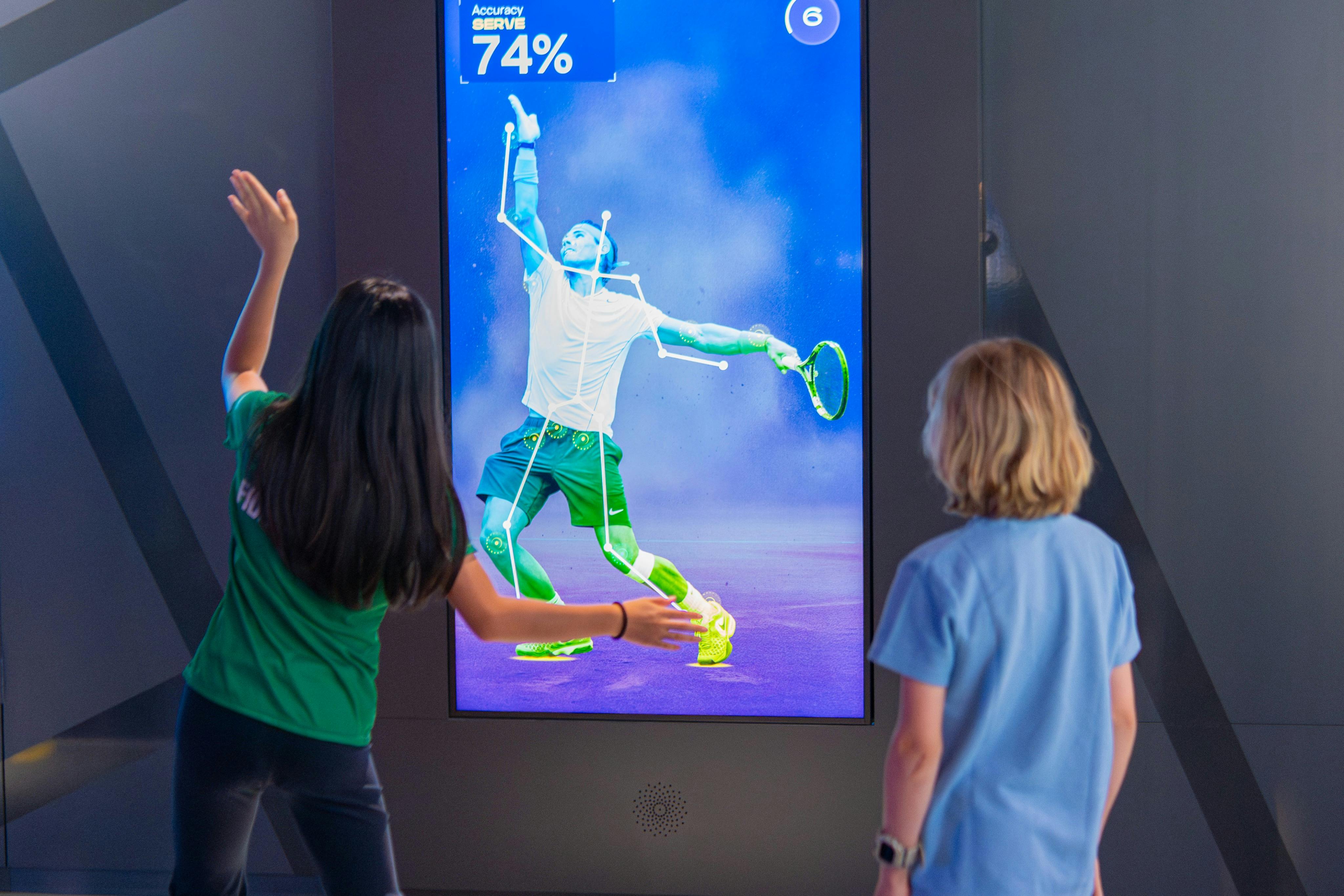 Two children interact with a digital display showing a tennis player with motion tracking and a 74% accuracy score.