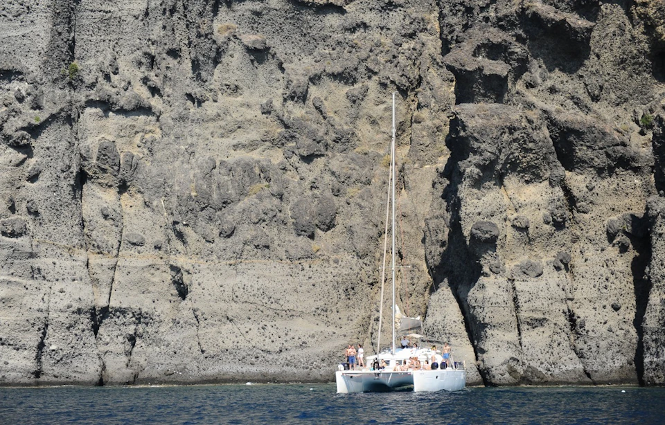 A catamaran with people on board floating near a large rocky cliff face.