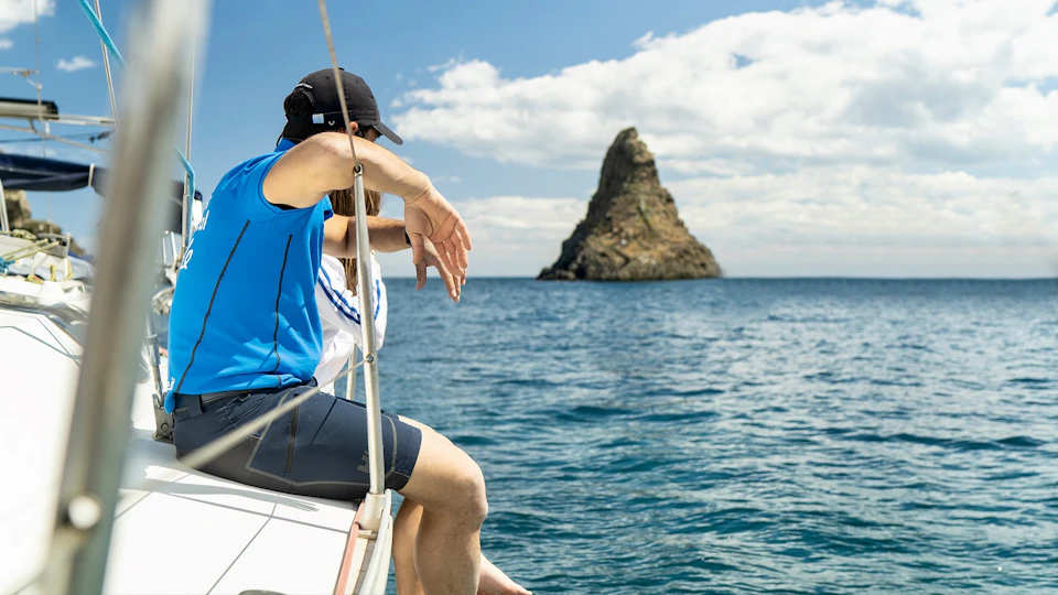 Two people sitting on a boat, looking at a distant rock formation in the ocean under a partly cloudy blue sky.