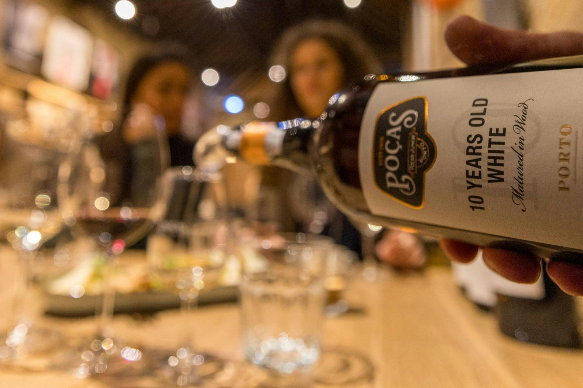 Een close-up van een fles met het label "10 Years Old White" die wordt ingeschonken, met vage mensen en een gedekte tafel op de achtergrond.