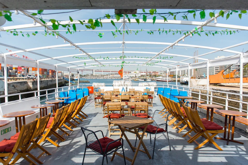 Open seating area on a boat deck with wooden chairs and tables, decorated with green vines, overlooking a harbor.