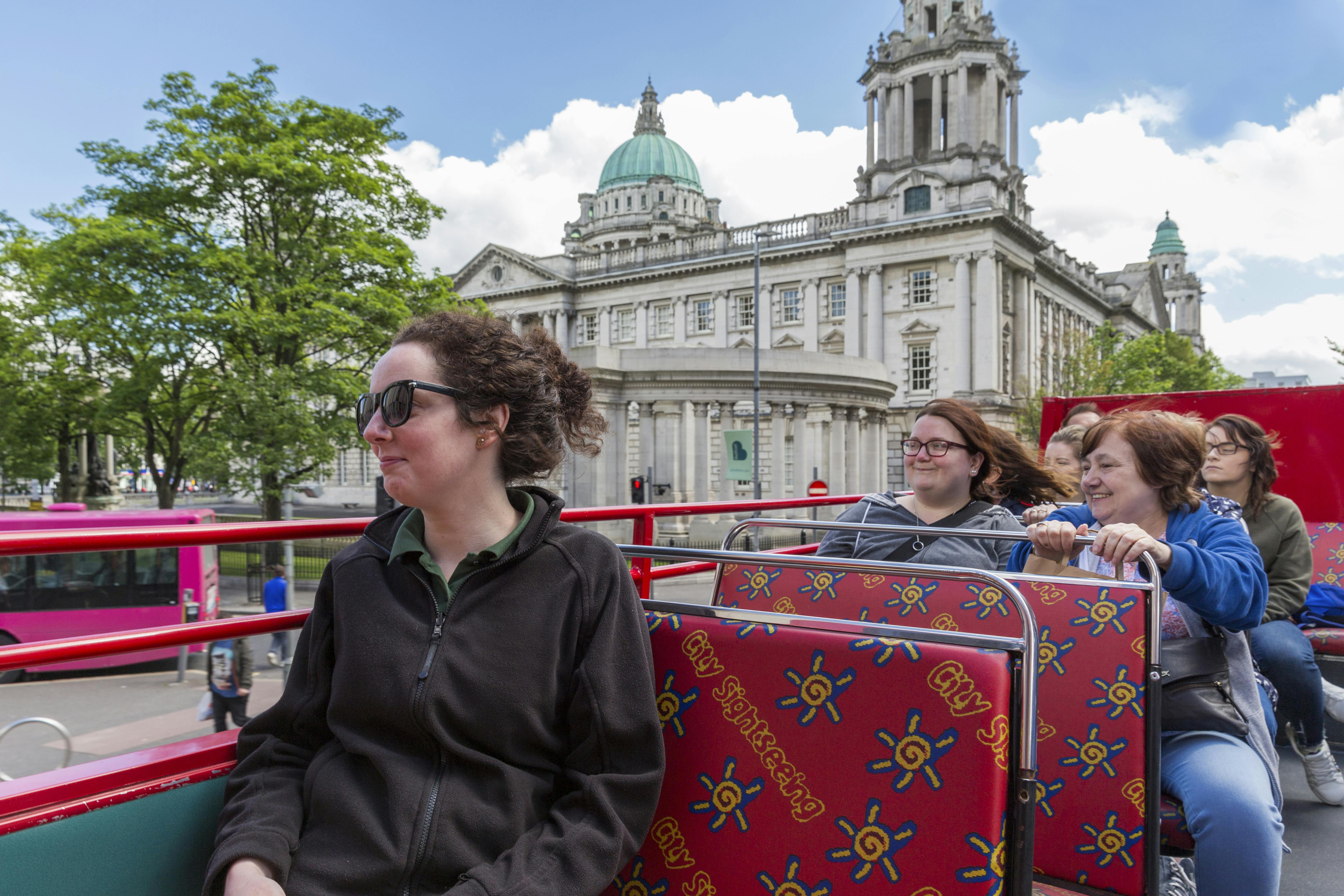 Turistas en un autobús turístico descubierto delante de un edificio histórico con cúpulas y columnas bajo un cielo parcialmente nublado.