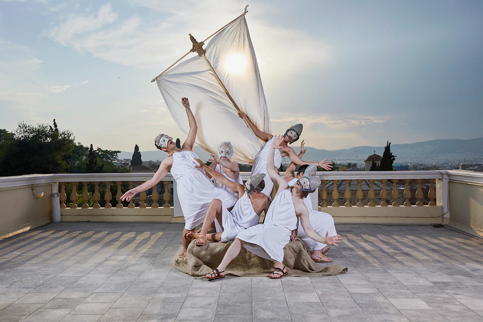 Six people in togas and masks pose dramatically with a makeshift sail against an outdoor backdrop and skyline.