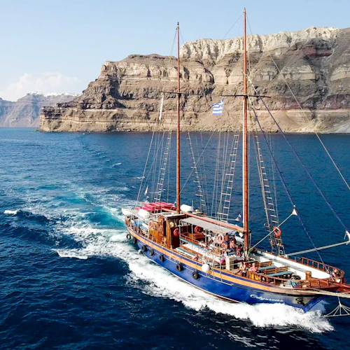 Ein großes Segelboot mit blauem Rumpf segelt in der Nähe von felsigen Klippen in klarem, blauem Wasser, und an Deck sind Menschen zu sehen.