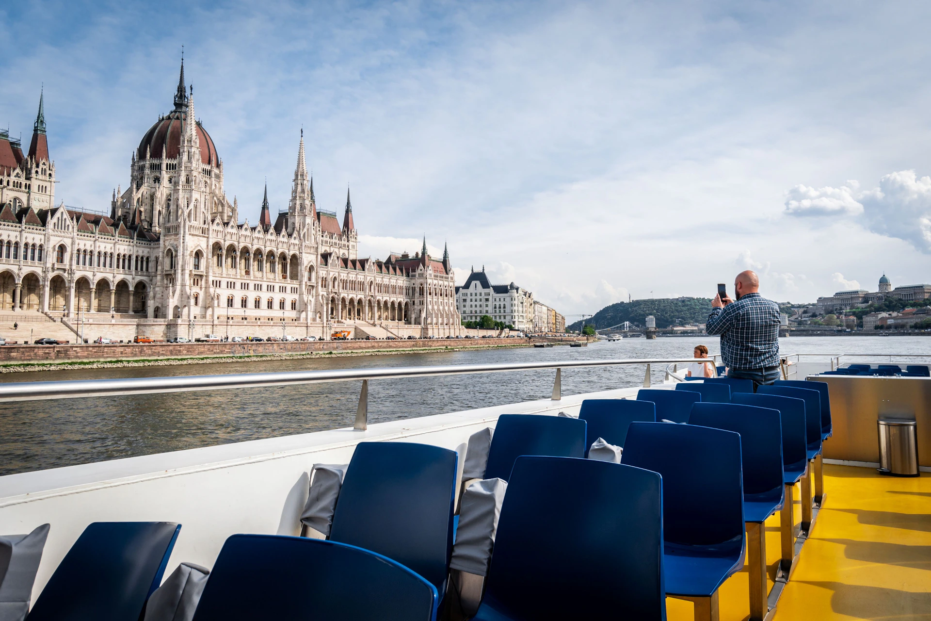 View of the Hungarian Parliament Building from a boat with empty blue chairs and a person taking a photo in the foreground.