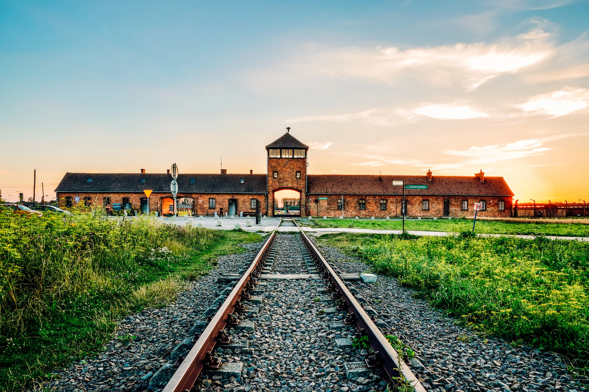 Railway tracks leading to a brick building with a central watchtower, surrounded by grassy fields under a partially cloudy sky.