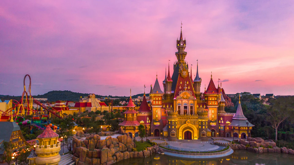 A colorful castle with spires at sunset, surrounded by landscaped greenery and water, under a purple-pink sky.