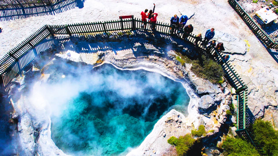 People standing on a wooden fence overlook a steaming, vibrant turquoise geothermal pool surrounded by rock formations.